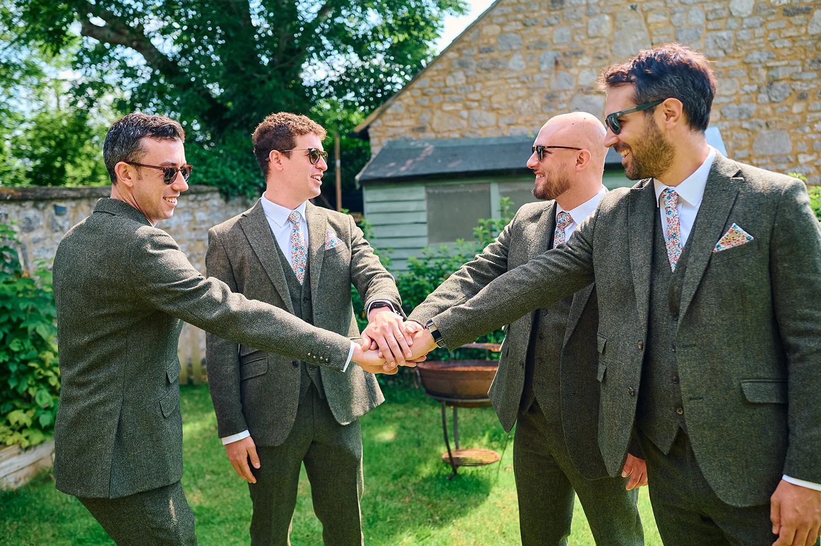Four groomsmen wearing matching green tweed suits, floral ties and sunglasses stack their hands together in a sunny garden setting.