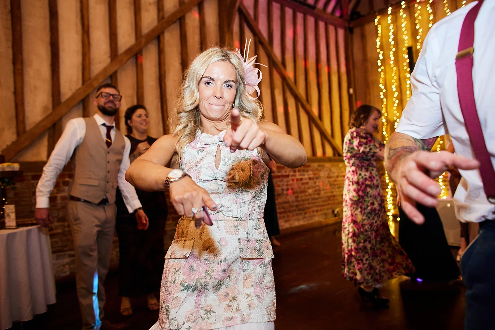 A wedding guest in a floral print dress and fascinator points playfully at the camera while dancing on a packed evening dance floor inside a rustic barn.