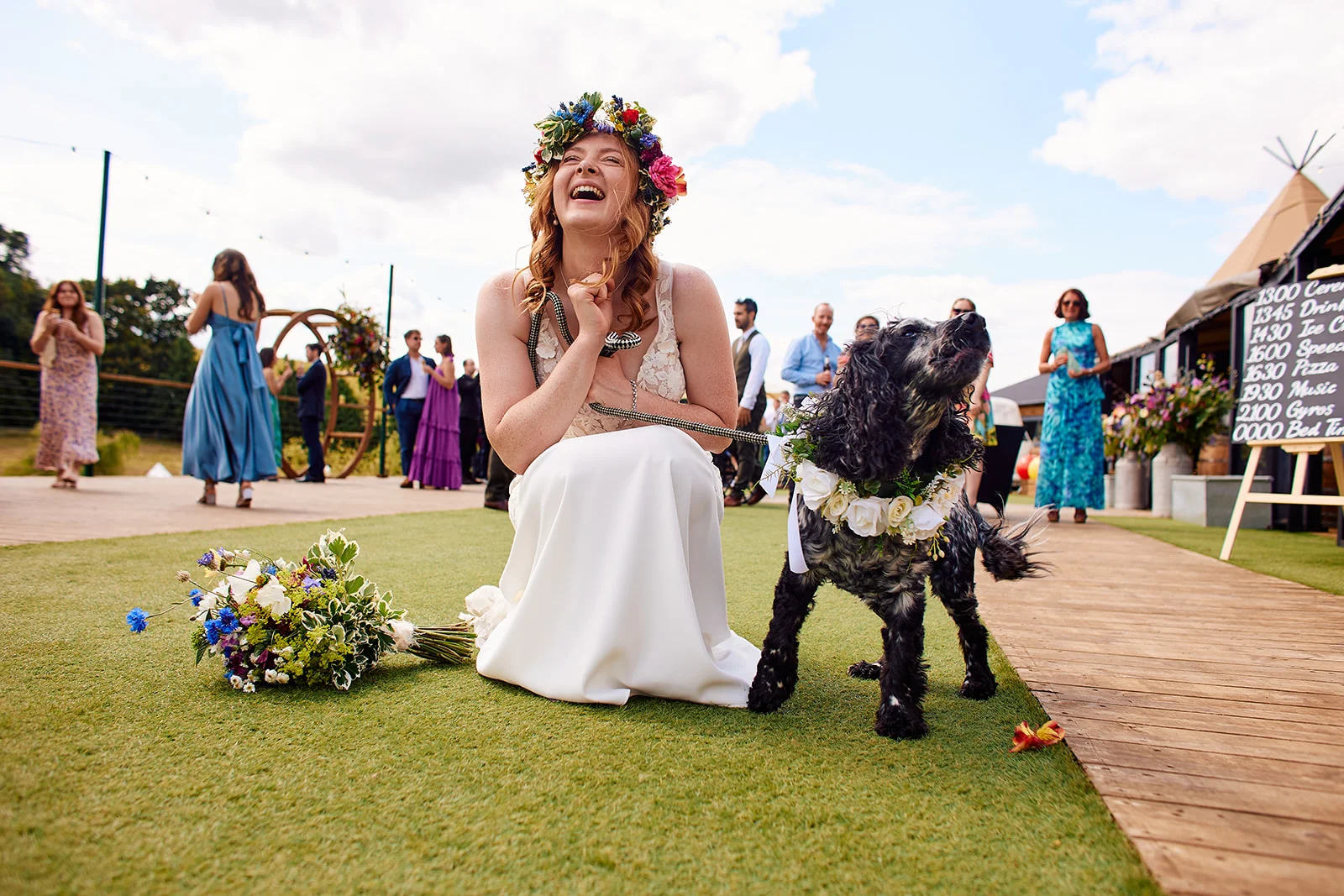 A joyous bride wearing a colourful flower crown kneels on the grass and laughs alongside her spaniel dog, who wears a matching floral collar during an outdoor wedding celebration.