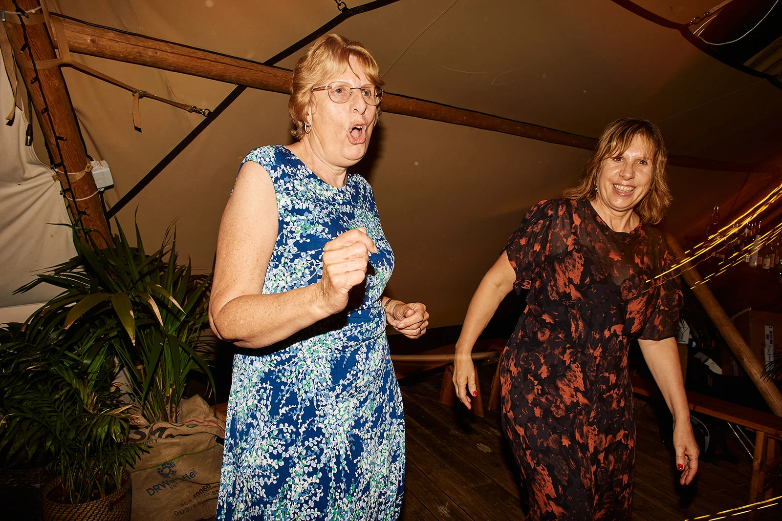 Two older female guests laugh and dance energetically inside a marquee wedding reception illuminated by direct flash.