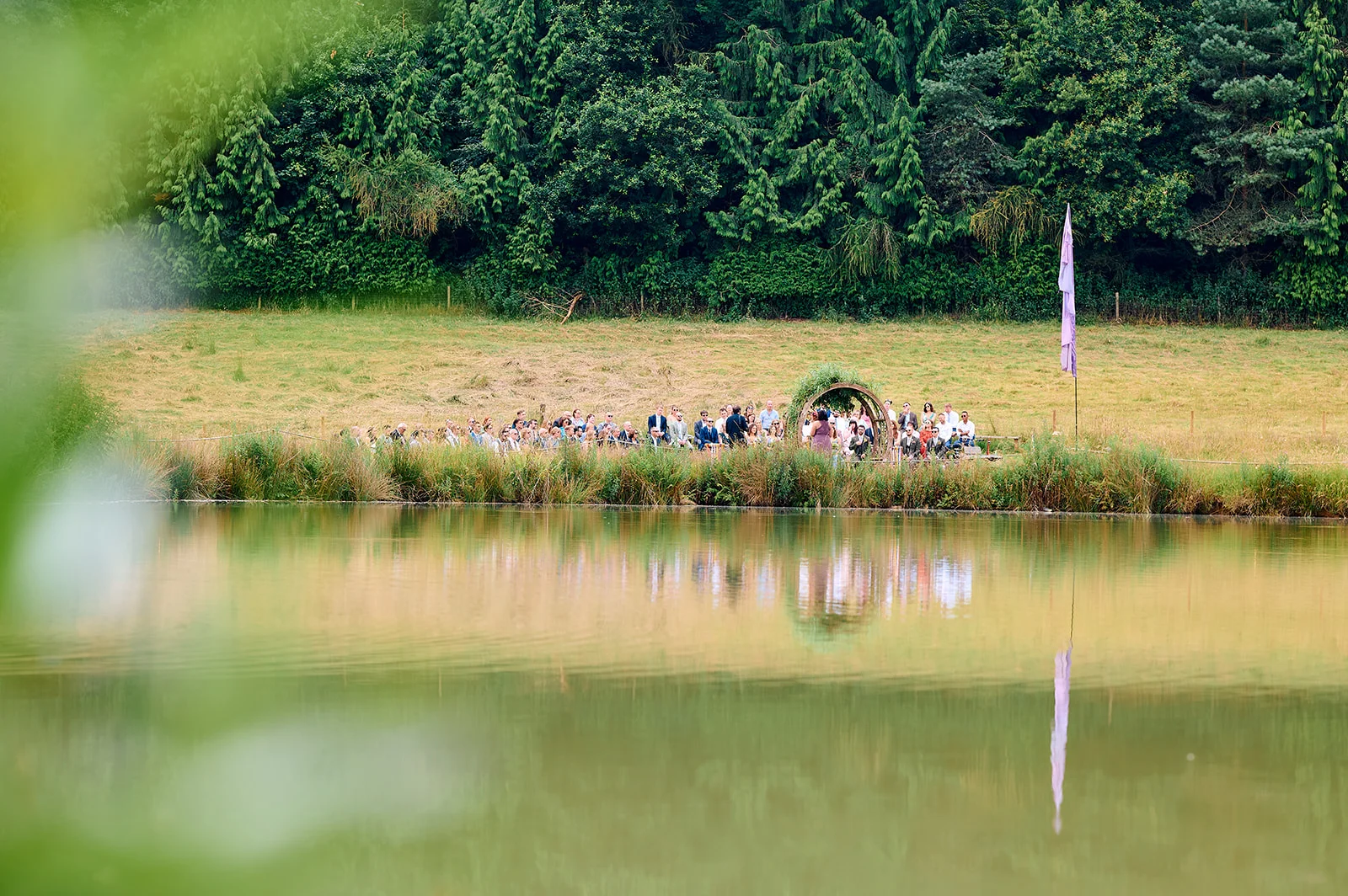An outdoor wedding ceremony takes place beside a tranquil lake with guests seated under a circular floral arch against a backdrop of dense woodland trees.