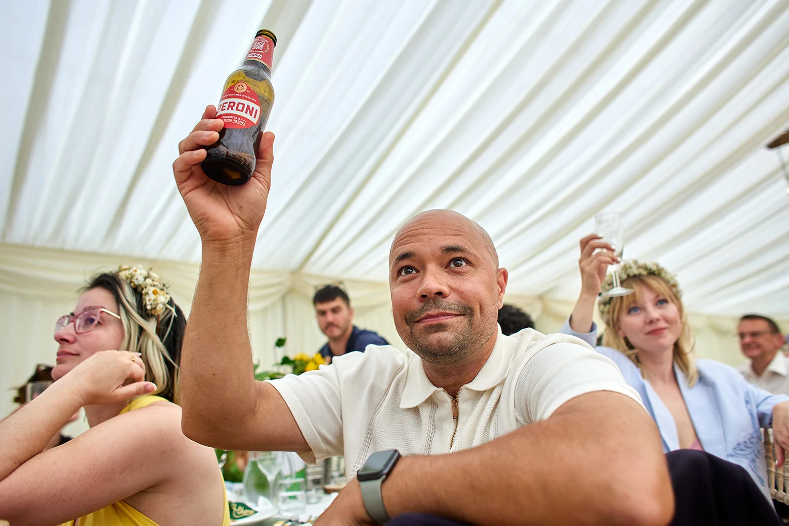 A joyful wedding guest cheerfully raises a bottle of beer in the air while seated inside a draped marquee reception.