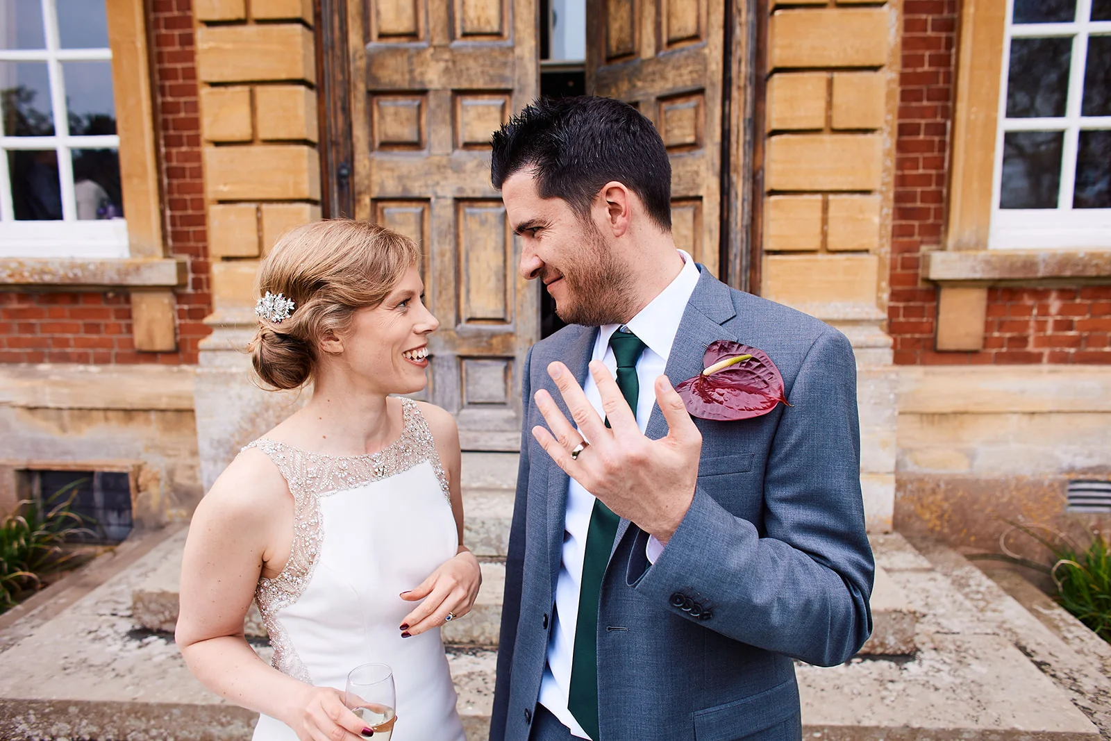 A smiling bride and groom share a joyful glance outside a grand stone building, with the groom proudly displaying his wedding band.