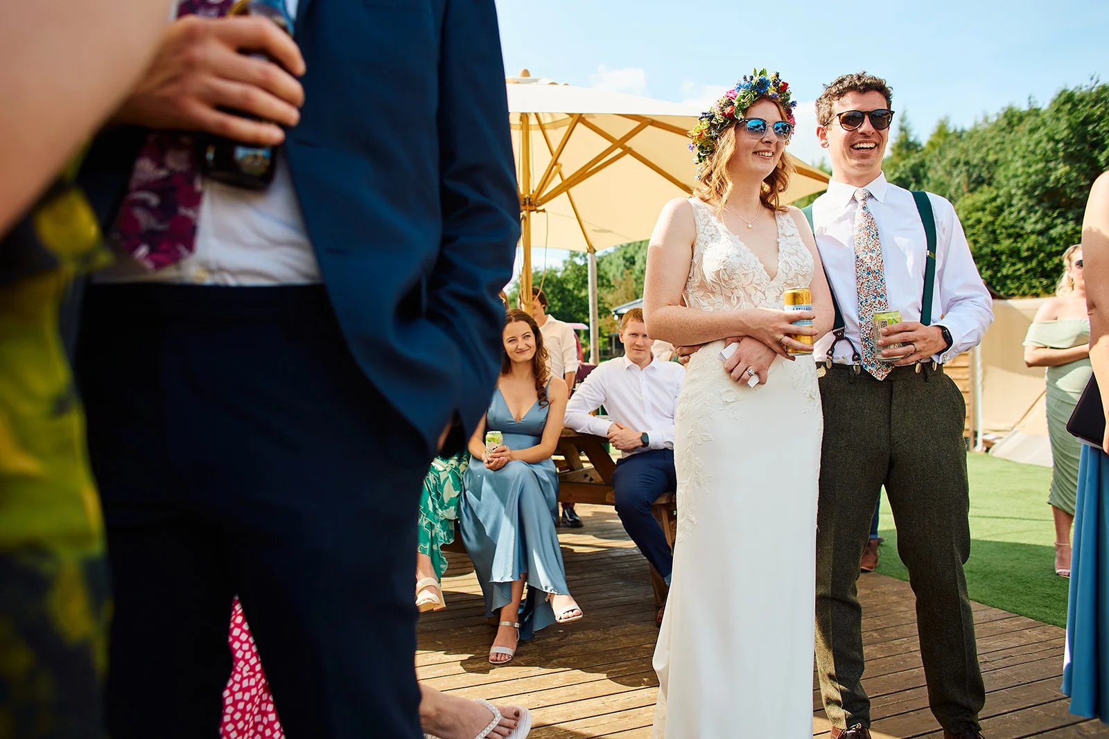A smiling bride wearing a floral crown and sunglasses stands with the groom in braces during a sunny outdoor wedding reception, with guests relaxing in the background.