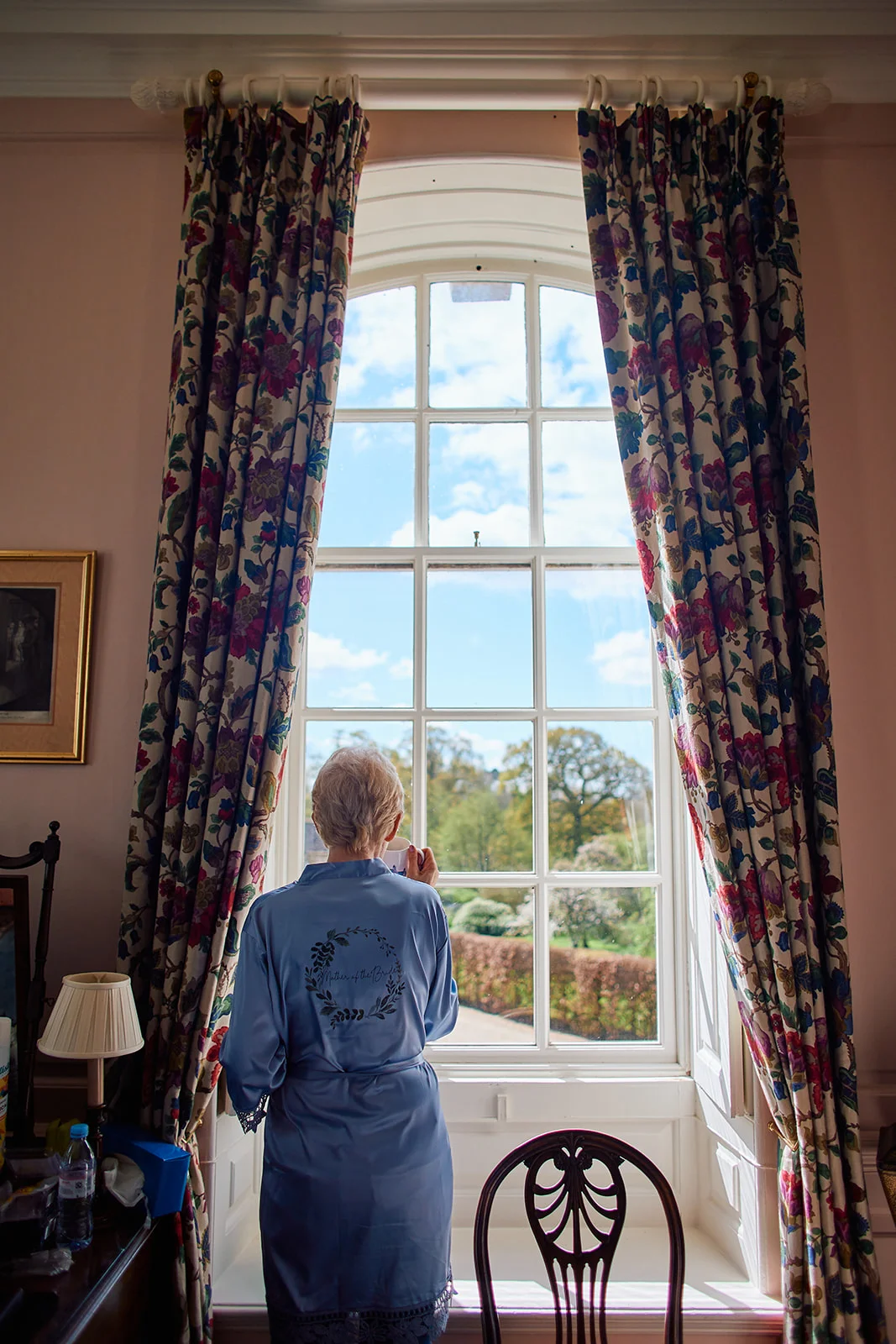 A mother of the bride stands by a tall arched window wearing a blue satin robe while looking out at the sunny day.