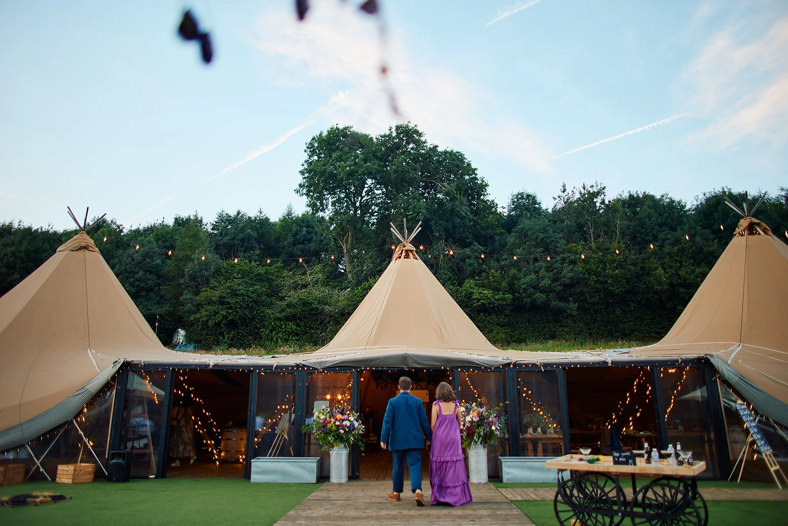 A couple walks hand in hand towards a connected cluster of illuminated tipis set against a backdrop of trees and a blue evening sky.