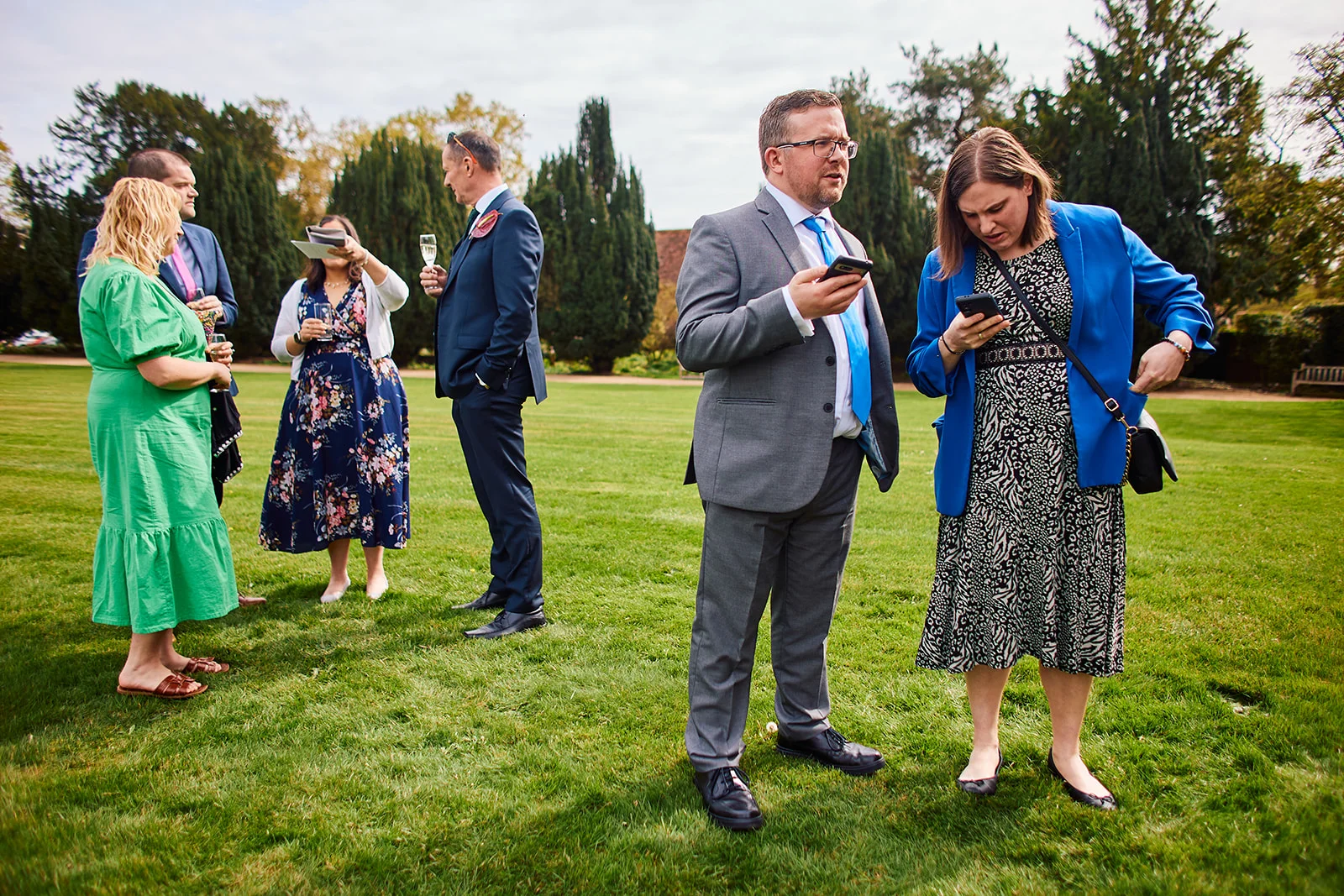 Wedding guests mingle and check their phones outdoors on a grassy lawn during the wedding reception.