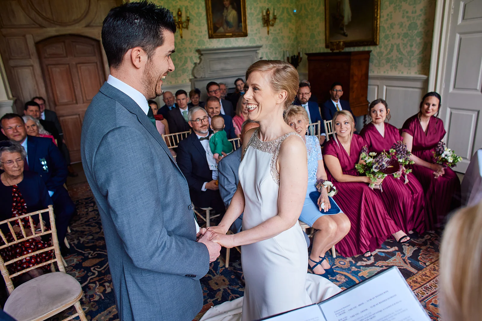 A smiling bride and groom hold hands and laugh together during their indoor civil ceremony in a historic panelled room, watched by seated guests and bridesmaids in burgundy dresses.