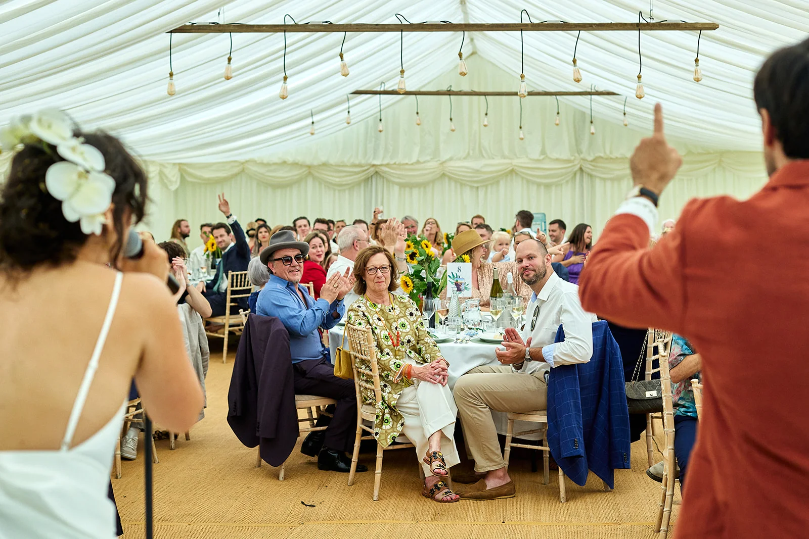 A guest delivers a speech during a wedding breakfast inside a marquee decorated with draped fabric and hanging lights.