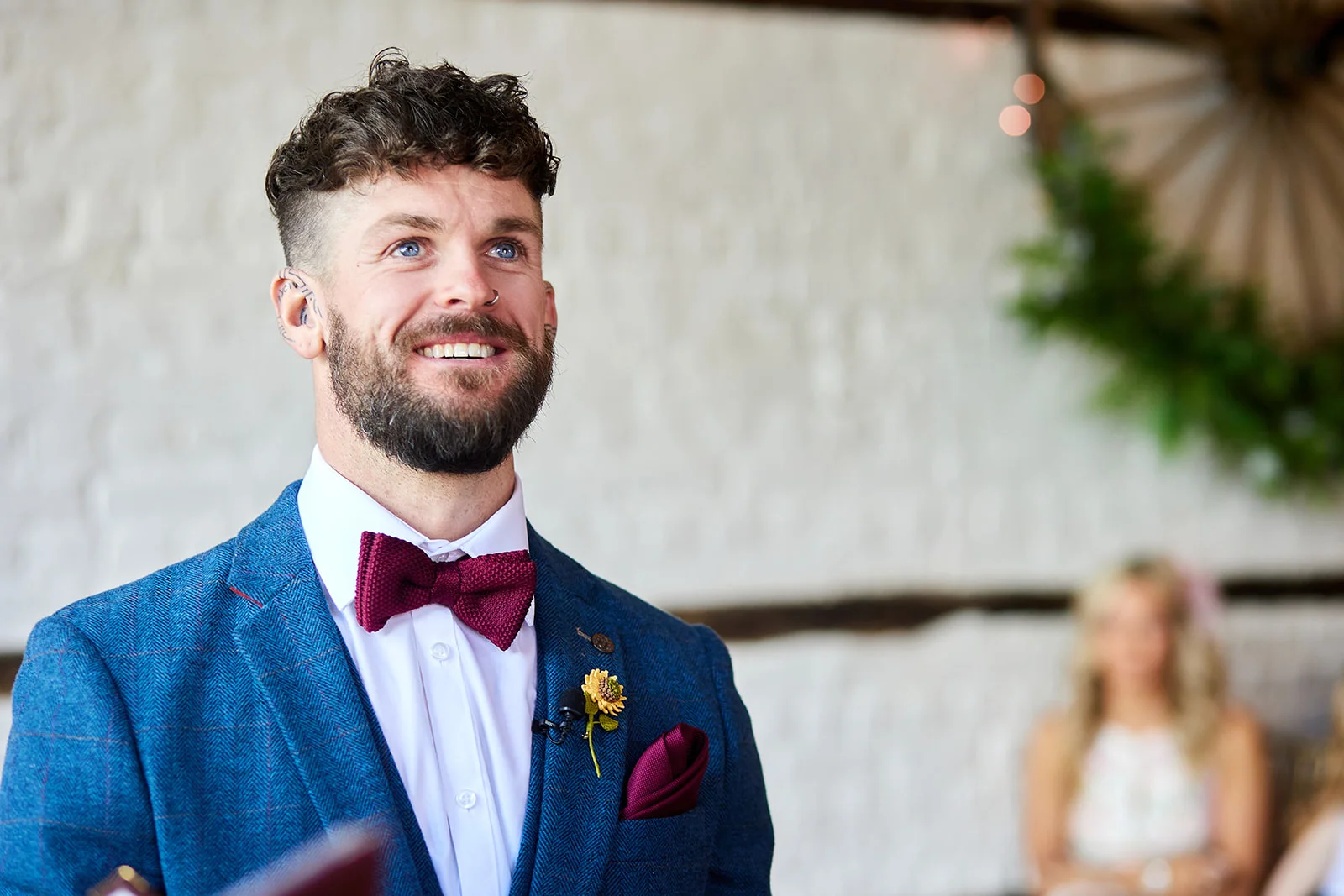 A smiling groom wearing a blue checked suit and a burgundy bow tie looks up emotionally during the wedding ceremony.