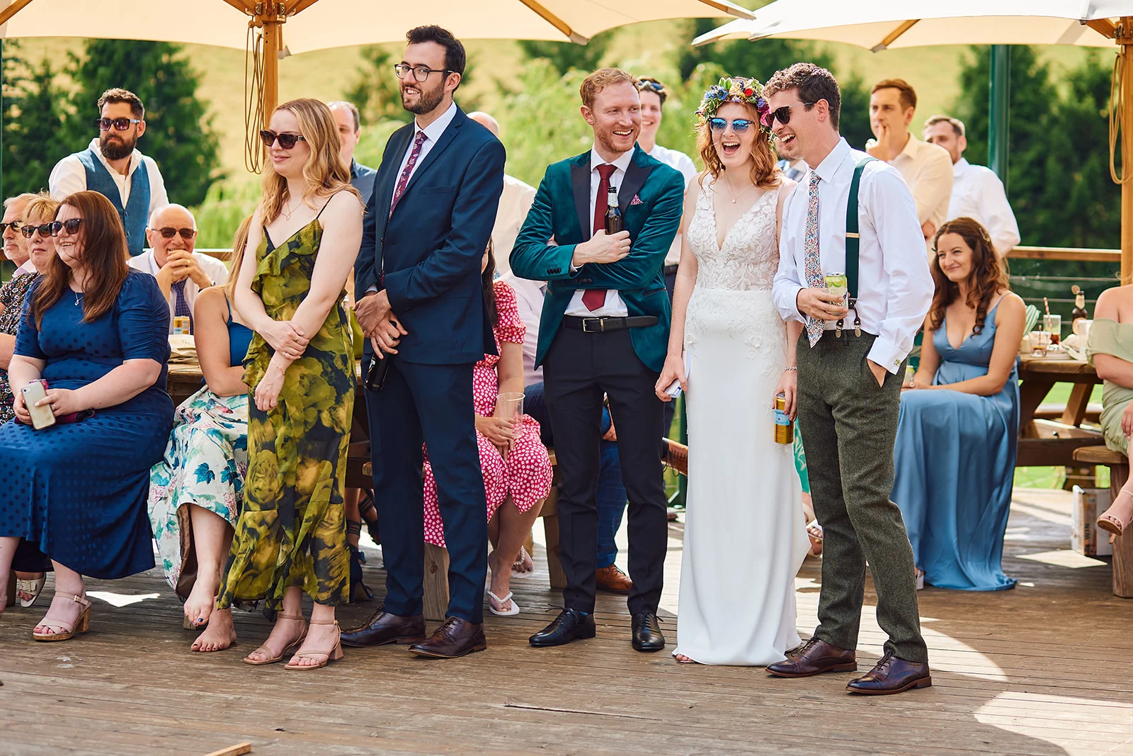 A joyful couple and wedding guests stand together on a wooden deck in the sunshine, wearing stylish attire, sunglasses, and laughing during an outdoor celebration.