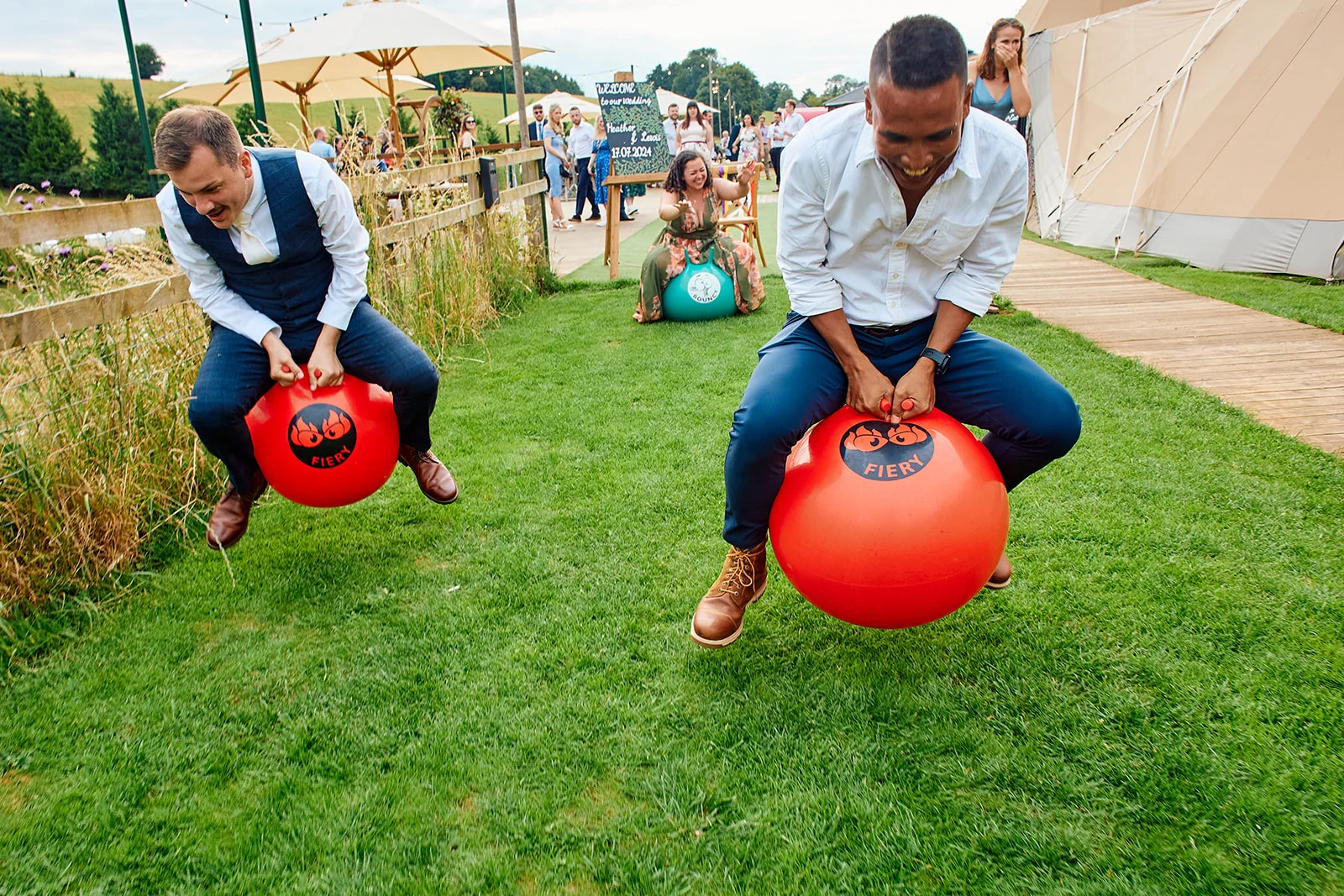 Two groomsmen laughing while bouncing on red space hoppers during an outdoor festival-style wedding reception.