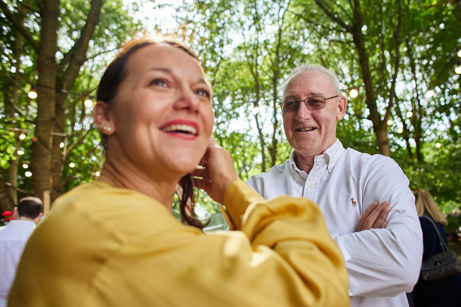 An outdoor wedding guest in a yellow top laughs happily while mingling with other guests under a canopy of trees.
