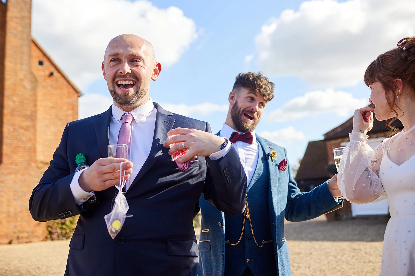 A joyful groom in a navy suit and pink tie laughs heartily during the outdoor drinks reception, accompanied by a laughing guest and bride under a clear blue sky.