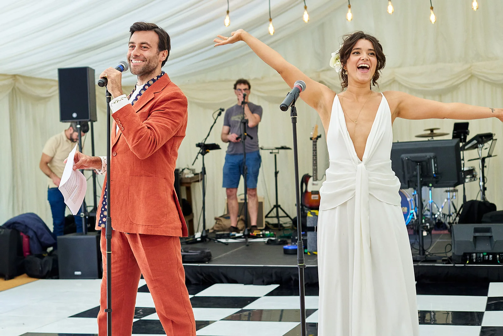 A joyful newlywed couple laughs and addresses guests into microphones inside a marquee with a live band performing on stage behind them.
