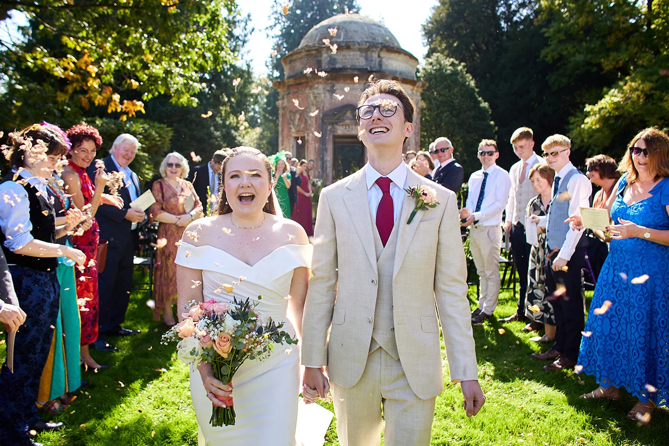 A joyful newlywed couple walks hand in hand through an outdoor ceremony as wedding guests shower them with celebratory confetti beneath a bright summer sky.