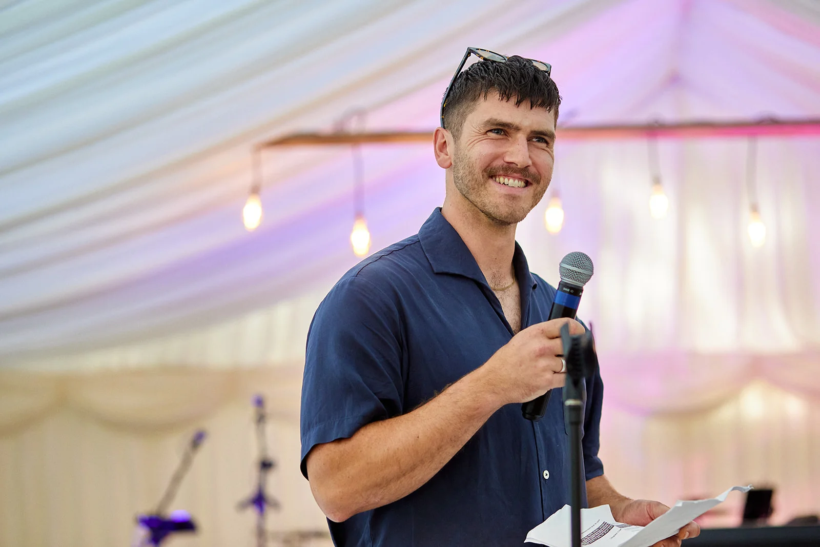 A smiling man holding a microphone and speech notes inside a marquee decorated with white ceiling drapes and hanging string lights.