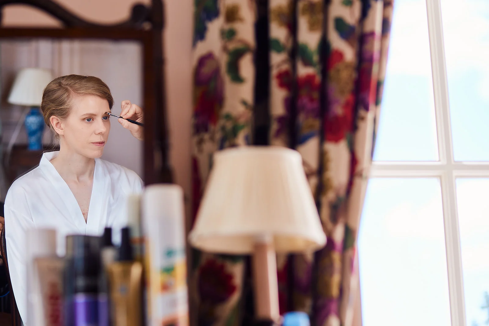 A bride sits calmly in a white robe while having her makeup applied in front of a mirror during morning bridal preparations.