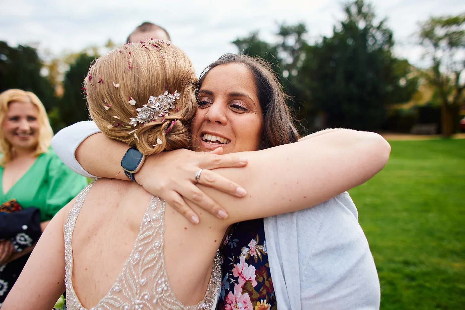 A guest and bride share a warm, joyful embrace outdoors in a garden setting, with confetti still caught in the bride's hair.
