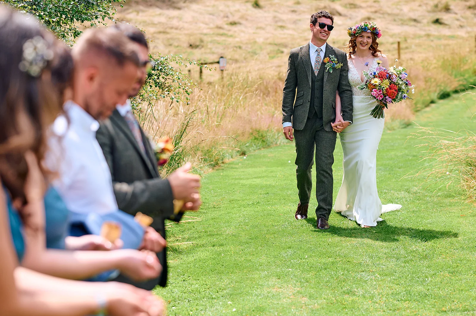 A newlywed couple smiles and holds hands while walking along a grassy path outdoors as guests cheer on the side.