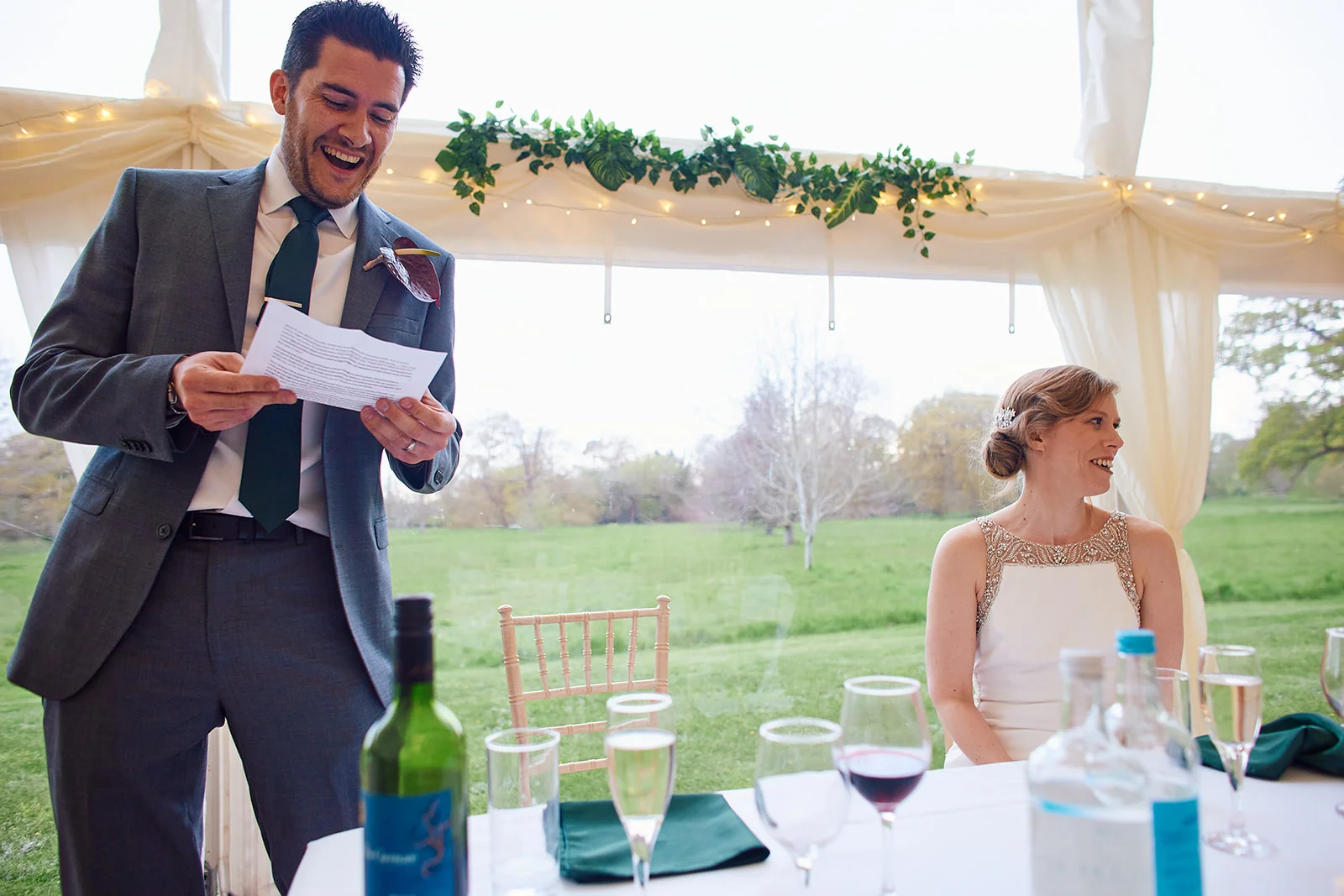 A groom in a grey suit laughs heartily while reading a speech inside a marquee, while the bride smiles nearby at a table set with wine glasses and greenery.