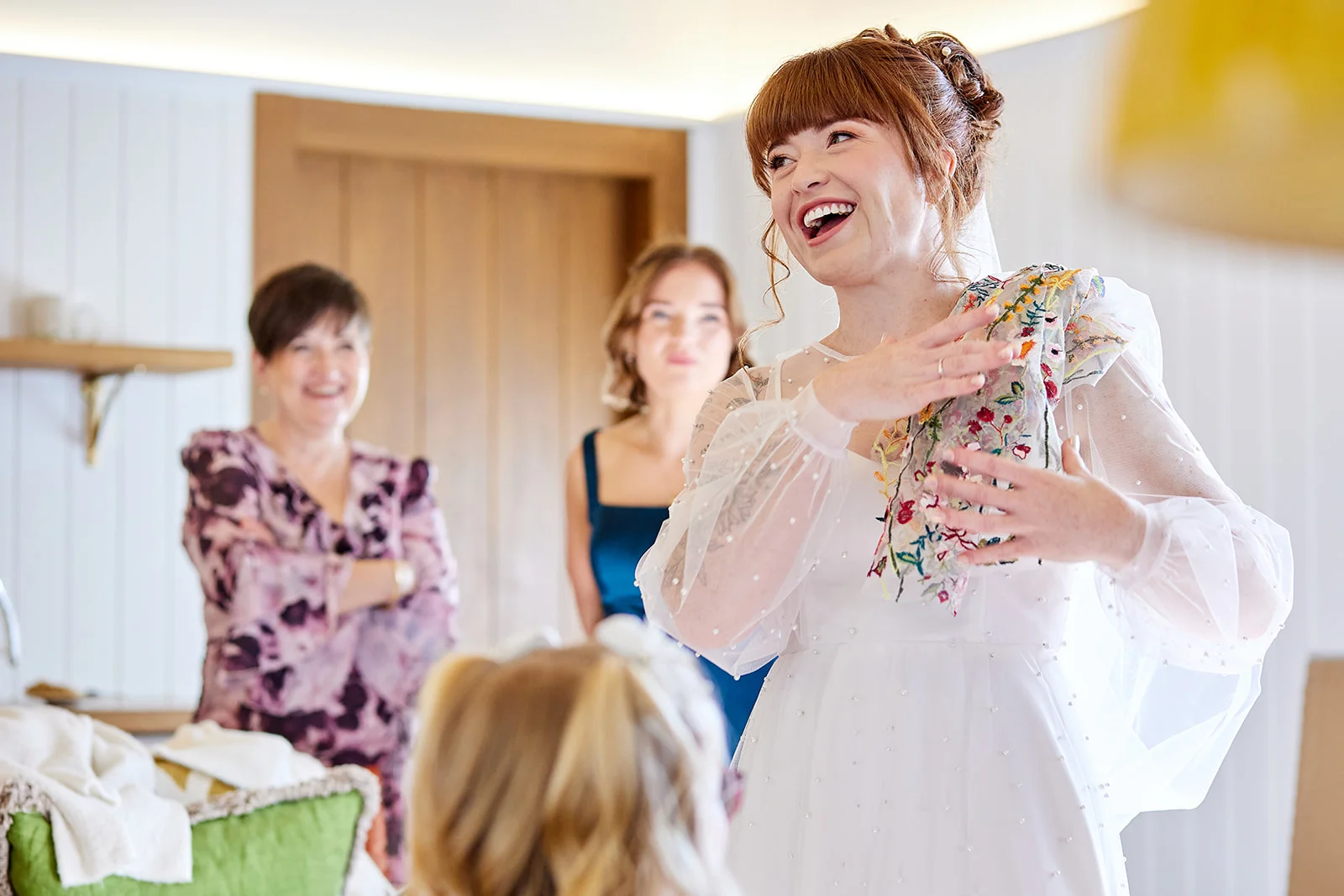 A joyful bride with red hair and a fringe laughs delightedly while trying on a sheer, embroidered long-sleeve wedding gown, surrounded by smiling family and bridesmaids during morning preparations.