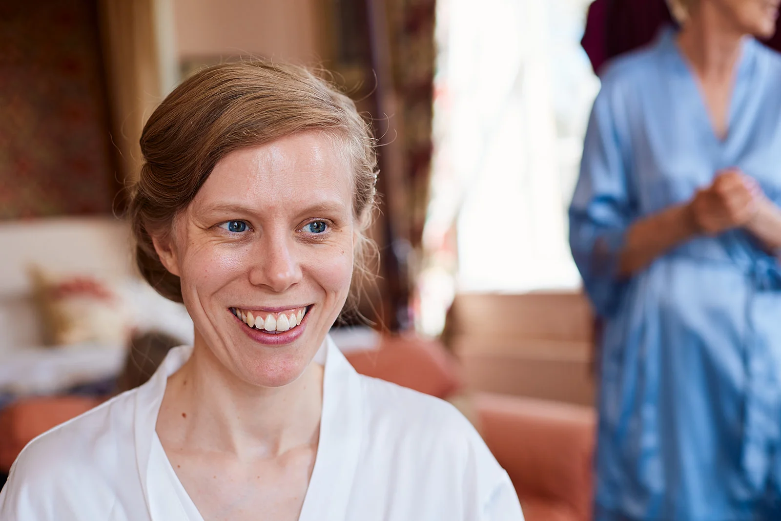 A smiling bride with blonde hair styled in a low chignon wears a white getting-ready robe while another person in a blue robe stands in the background.