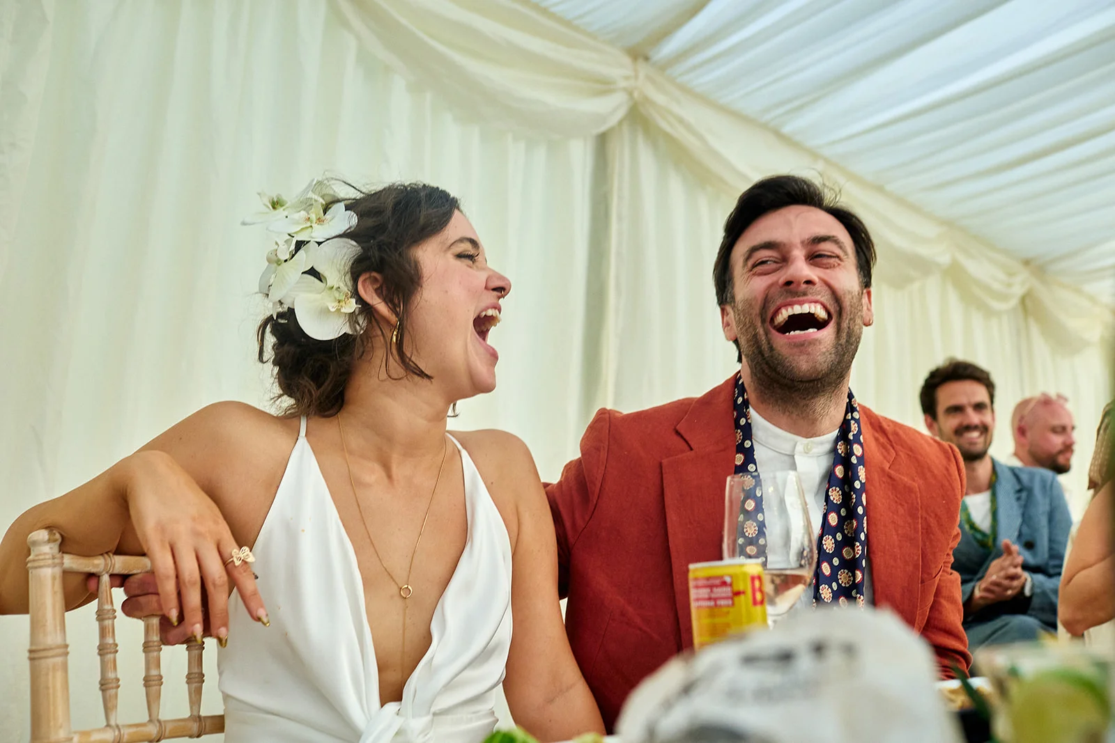 A joyful bride with white orchids in her hair and a groom in a rust-coloured jacket share a loud belly laugh seated inside a marquee during wedding speeches.