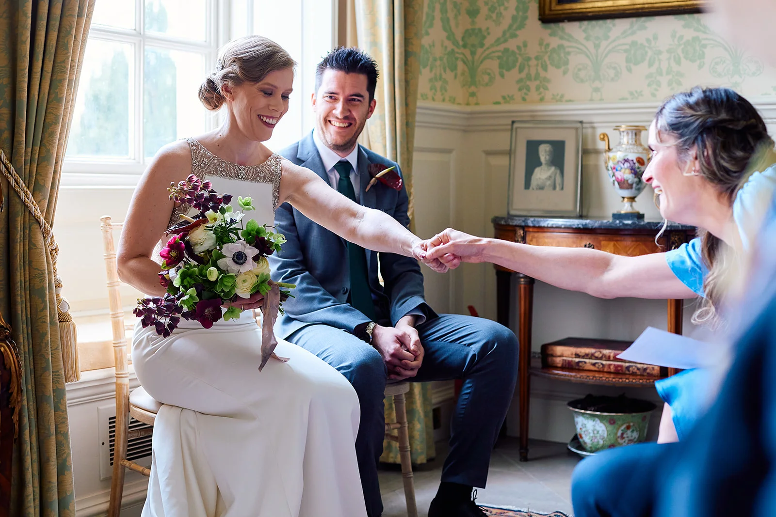 A smiling bride holding a bouquet of flowers sits beside her groom while greeting a guest inside a light-filled country house venue.