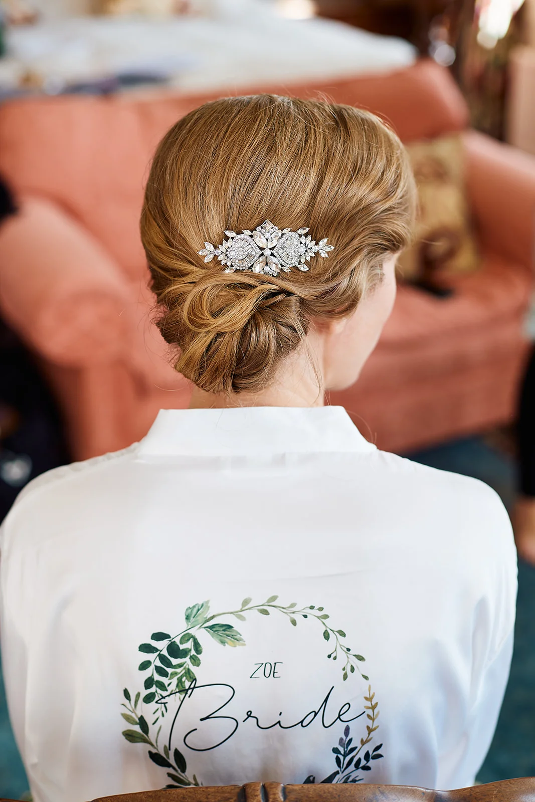 A bride viewed from behind showcases her intricate low bun hairstyle adorned with a sparkling crystal hair comb, while wearing a white personalised getting ready robe with a green botanical wreath design.