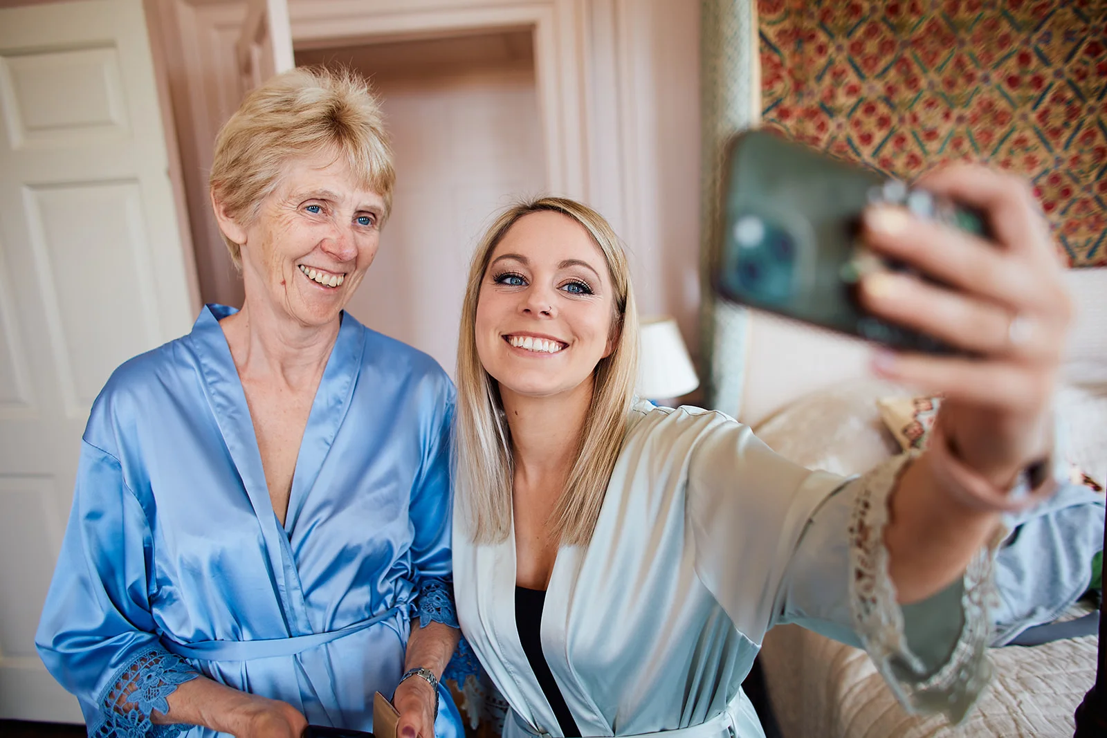 A smiling bride taking a selfie on her phone alongside her mother while both wear satin getting ready robes indoors.