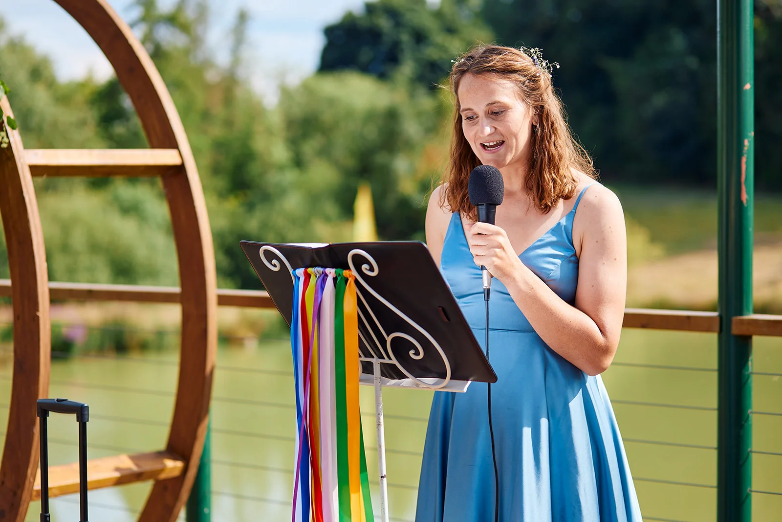A woman delivers a reading into a microphone outdoors during a sunny wedding ceremony, standing beside a decorated music stand.