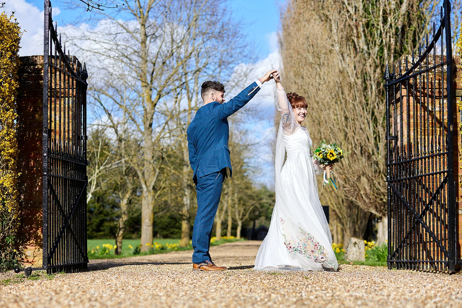 A newlywed couple joyfully twirls together on a gravel driveway framed by large black wrought iron gates and tall trees on a sunny spring day.