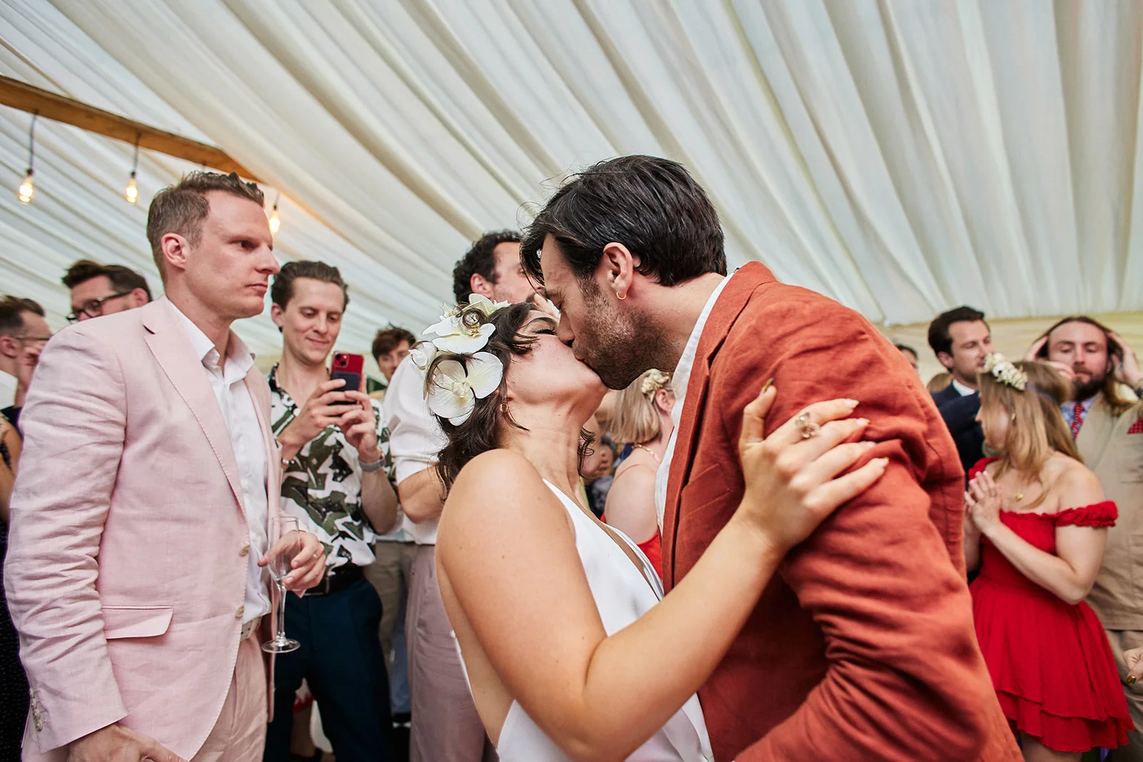 A newlywed couple share an intimate kiss in a marquee surrounded by cheering wedding guests, with the groom wearing a rust-coloured suit and the bride wearing white with white flowers in her hair.