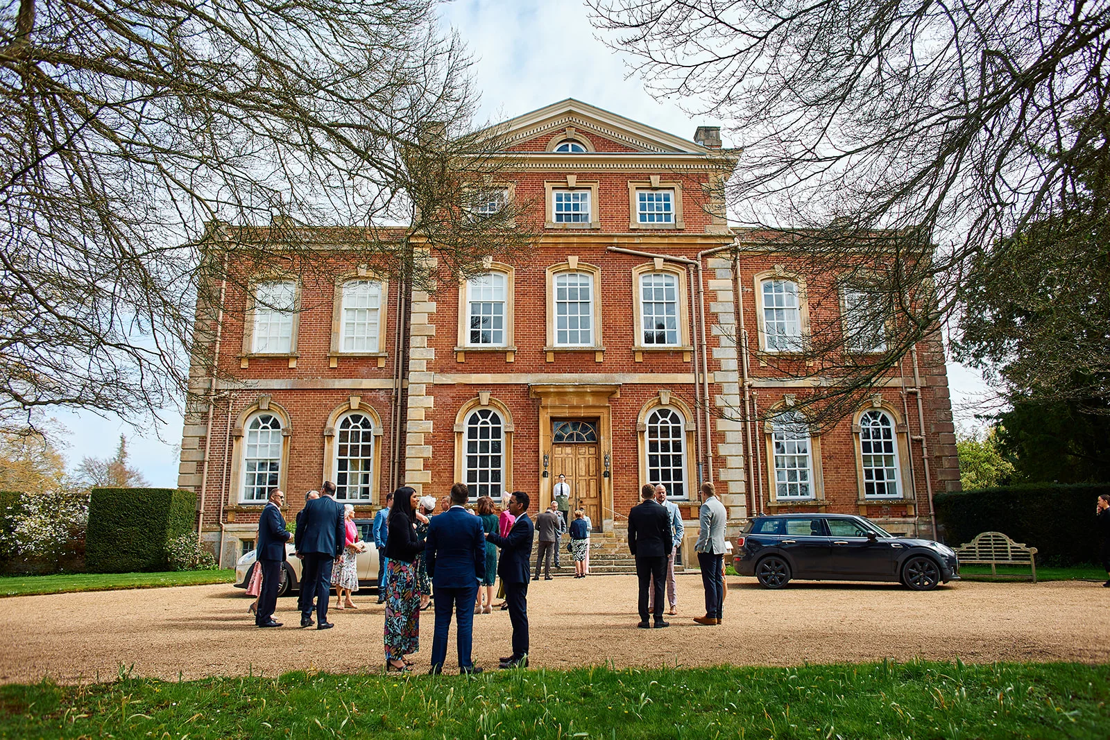 Wedding guests mingle outside a grand red brick Georgian manor house framed by bare tree branches on a bright day.