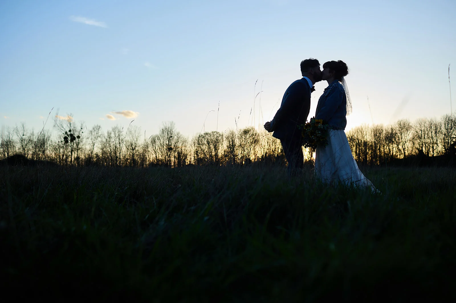 A newlywed couple shares a tender kiss in silhouette against a golden sunset sky while standing in a grassy field.