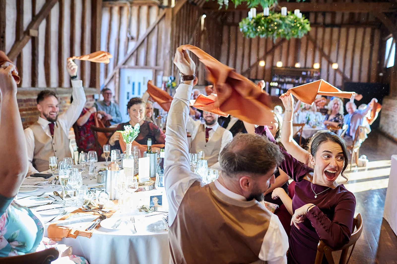Wedding guests enthusiastically wave orange napkins in the air during a lively reception inside a rustic oak-framed barn.