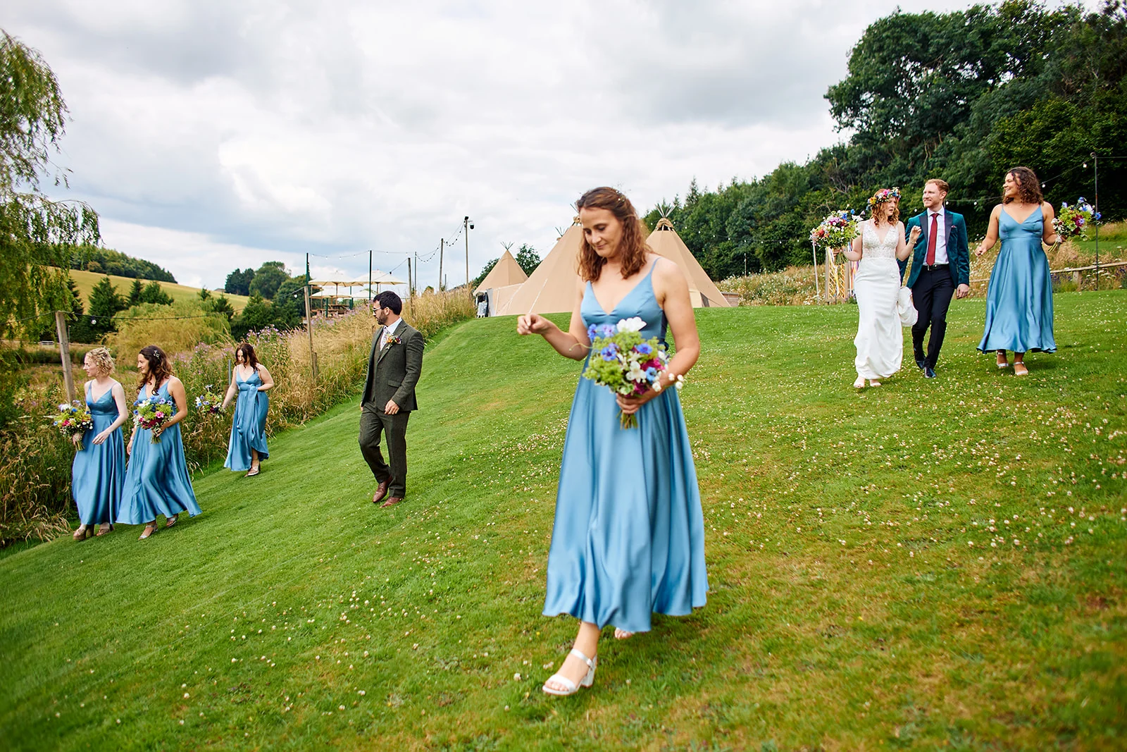 A bridal party in dusty blue dresses and a groom in a green suit walk across a grassy meadow towards tipis at an outdoor festival-style summer wedding.