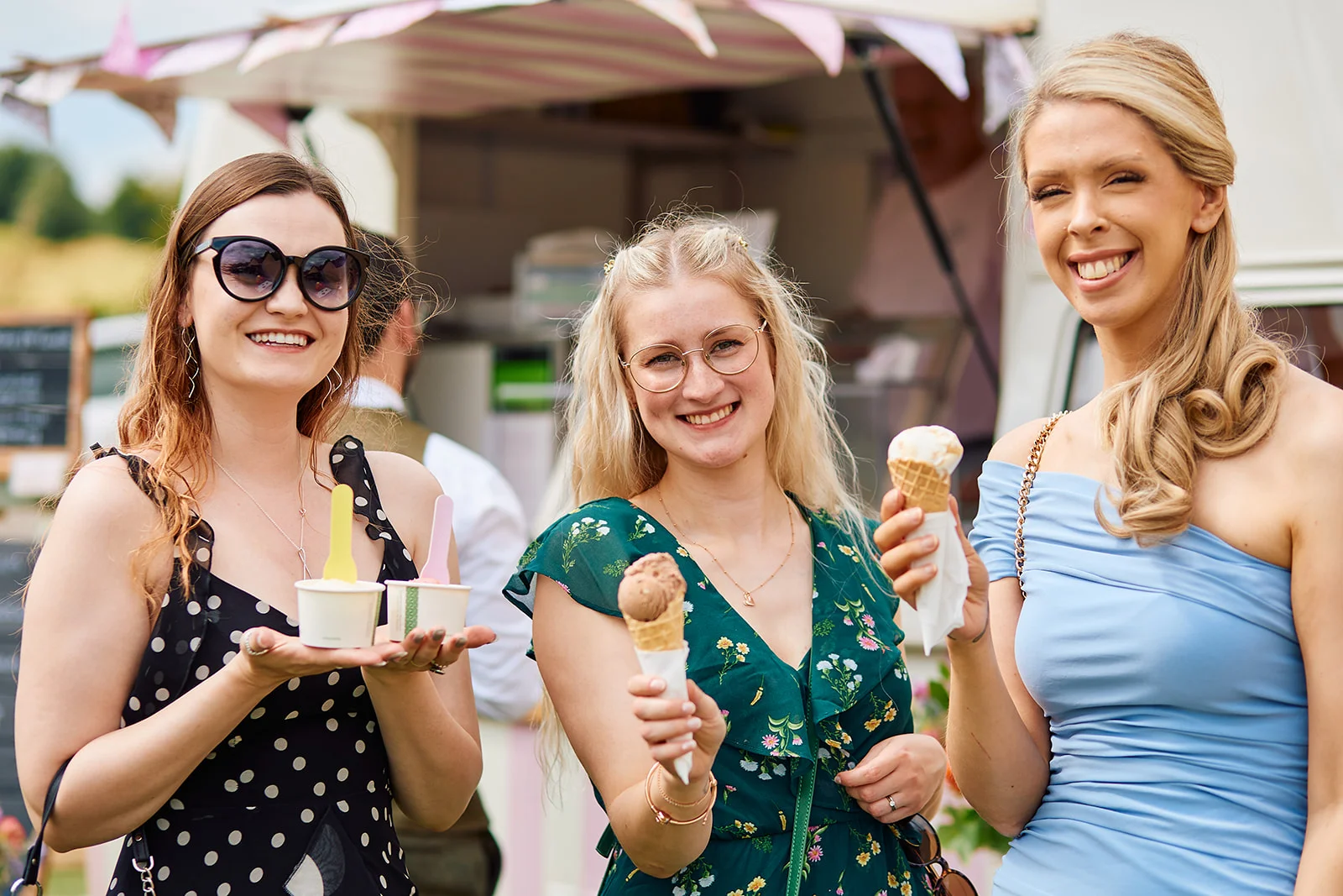 Three wedding guests joyfully holding ice creams and cones outside in front of a vintage ice cream van on a sunny summer day.