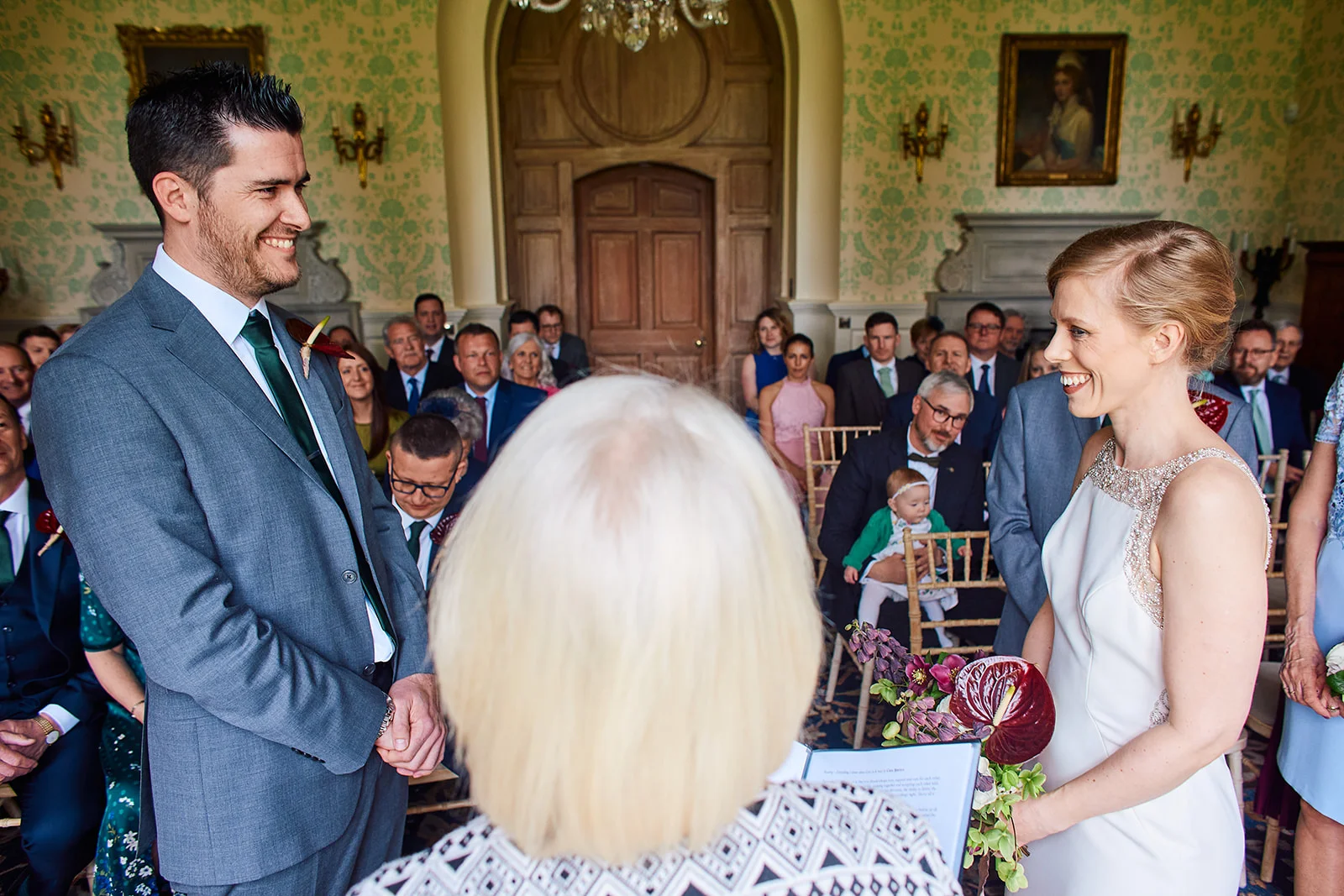 A smiling bride and groom stand facing each other during their civil ceremony inside an elegant wood-panelled room filled with seated wedding guests.