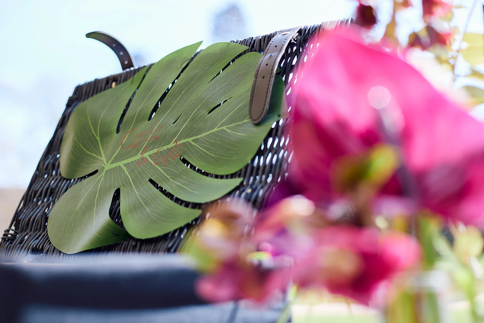 A close-up shot of a dark wicker basket featuring a large monstera leaf sign with handwritten calligraphy, framed by blurred vibrant pink tropical flowers in the foreground.