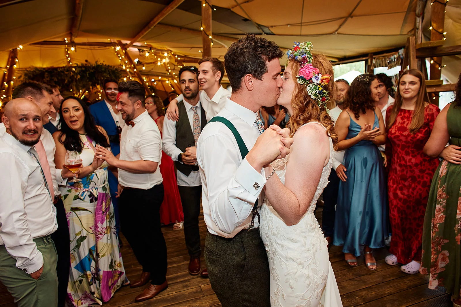 A newlywed couple shares a romantic kiss on the dance floor surrounded by cheering guests inside a festoon-lit marquee.