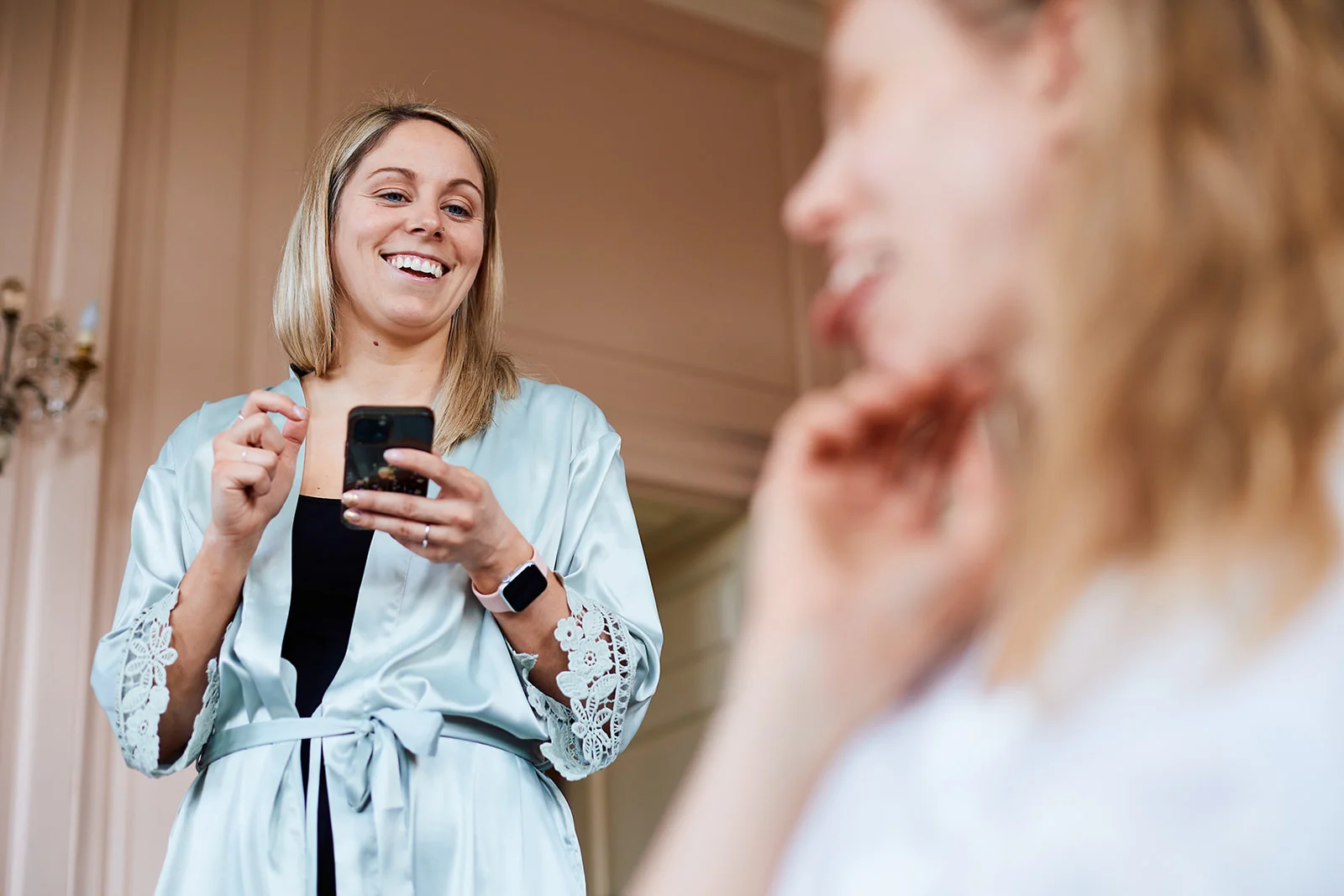A smiling bridesmaid wearing a light blue satin robe holds her smartphone during morning wedding preparations.
