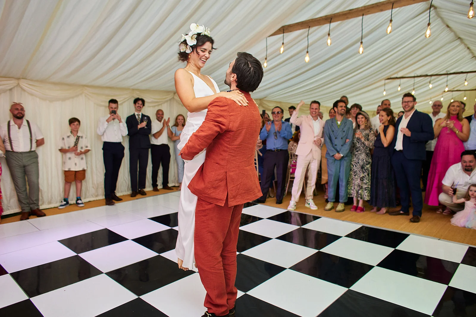 A groom lifts his laughing bride into the air on a black and white checkerboard dance floor inside a marquee filled with cheering wedding guests.