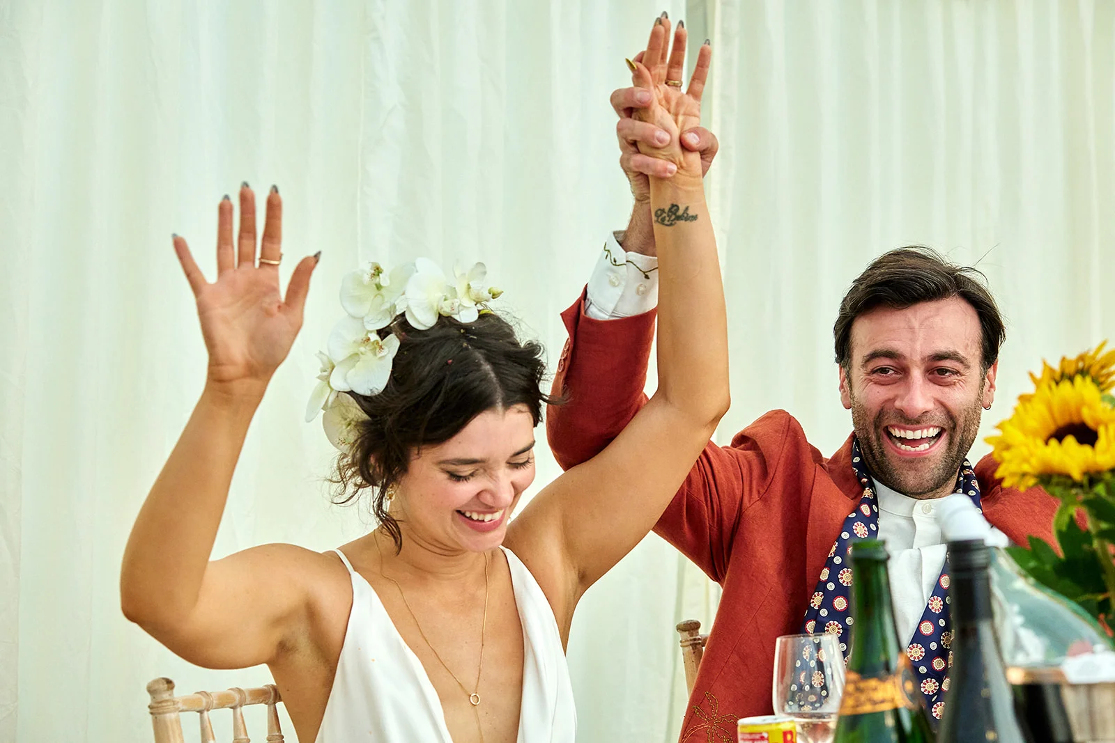 A joyful newlywed couple seated at the top table inside a marquee, laughing loudly with their hands raised high in celebration.