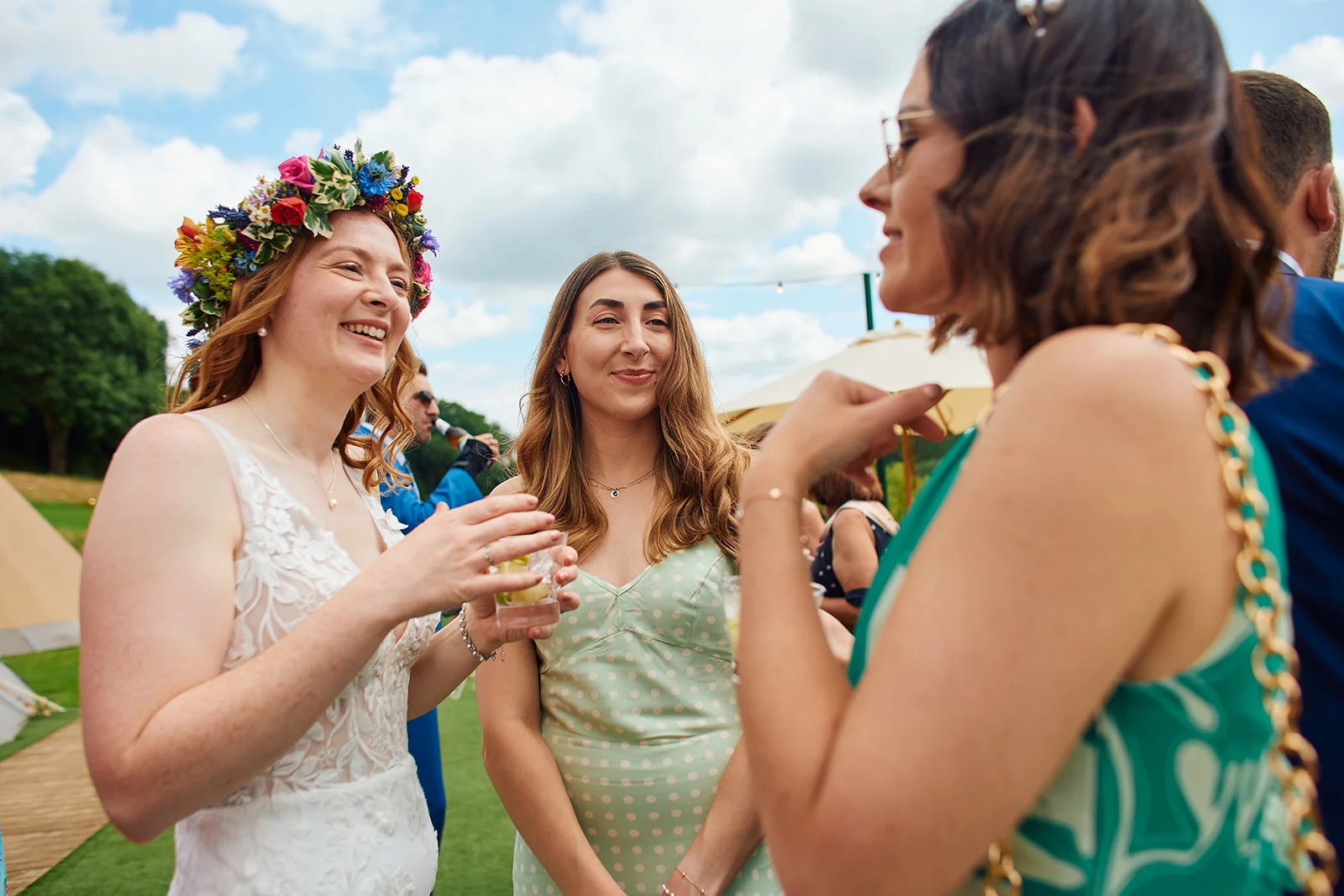 A smiling bride wearing a vibrant floral crown and lace gown chats with guests outdoors during a sunny wedding drinks reception.