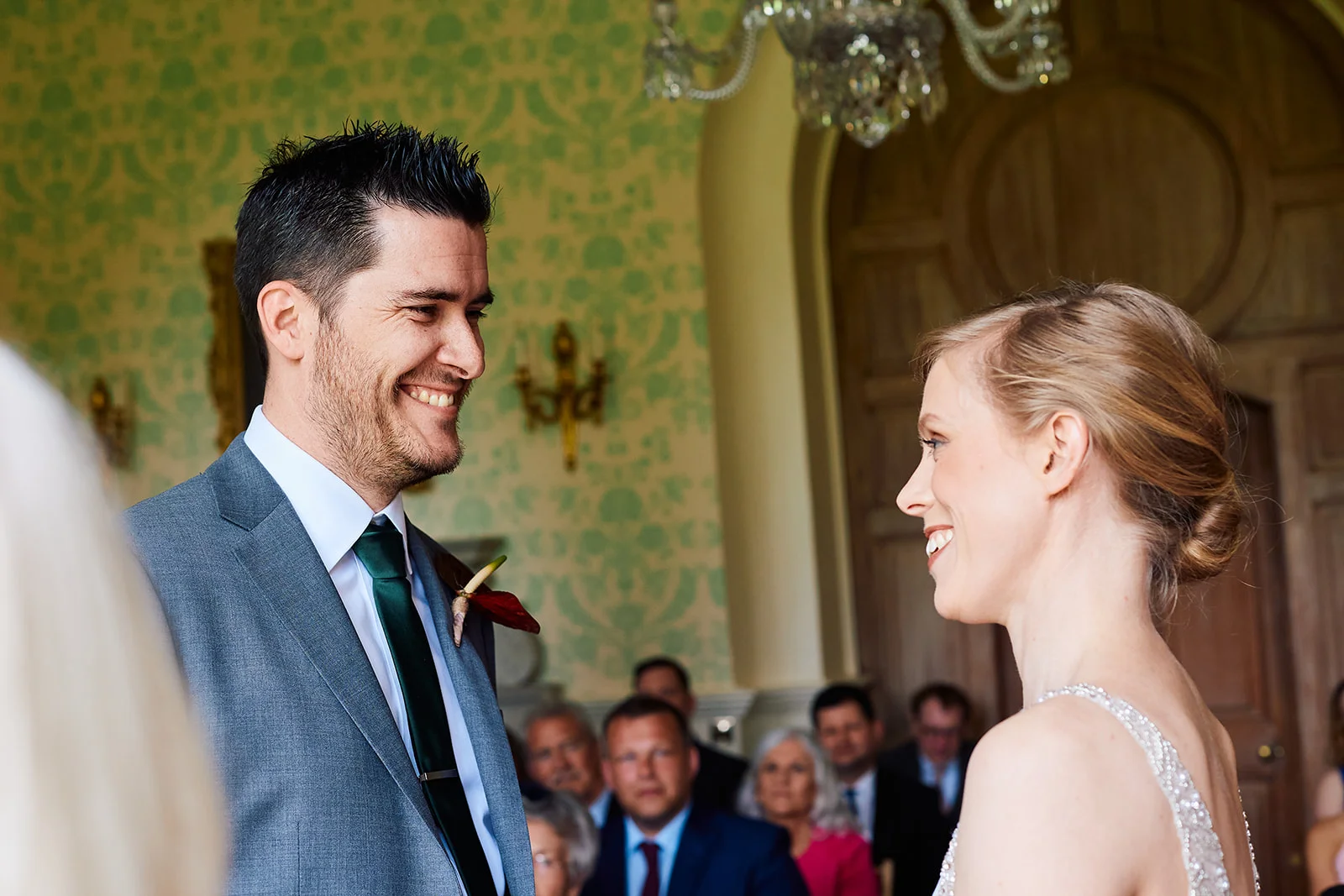 A joyful groom in a grey suit and green tie looks smilingly at his bride during their wedding ceremony inside a grand historic room.