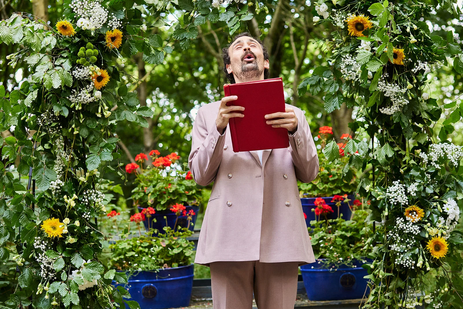 An emotional celebrant stands beneath a lush floral ceremony arch adorned with sunflowers and foliage, holding a bright red book with his eyes closed.