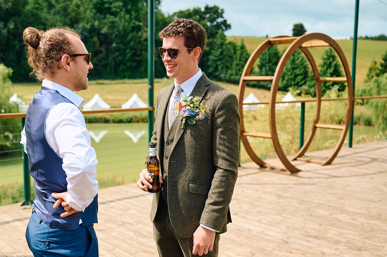 The groom in a green tweed suit and sunglasses chats with a guest holding a beer bottle outdoors near a circular wooden arch.