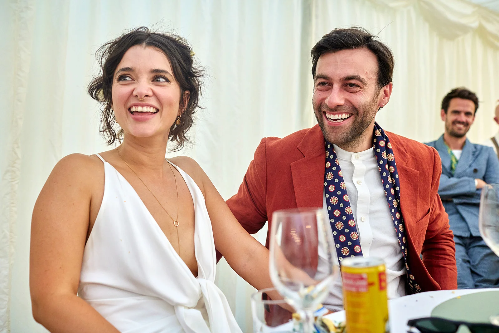 A smiling bride in a white slip dress and groom in a rust-coloured jacket laughing joyfully together at the wedding reception top table inside a marquee.